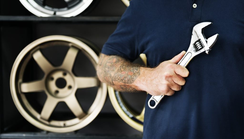A mechanic in a workshop holding a wrench with tyre/ wheel rims in the background